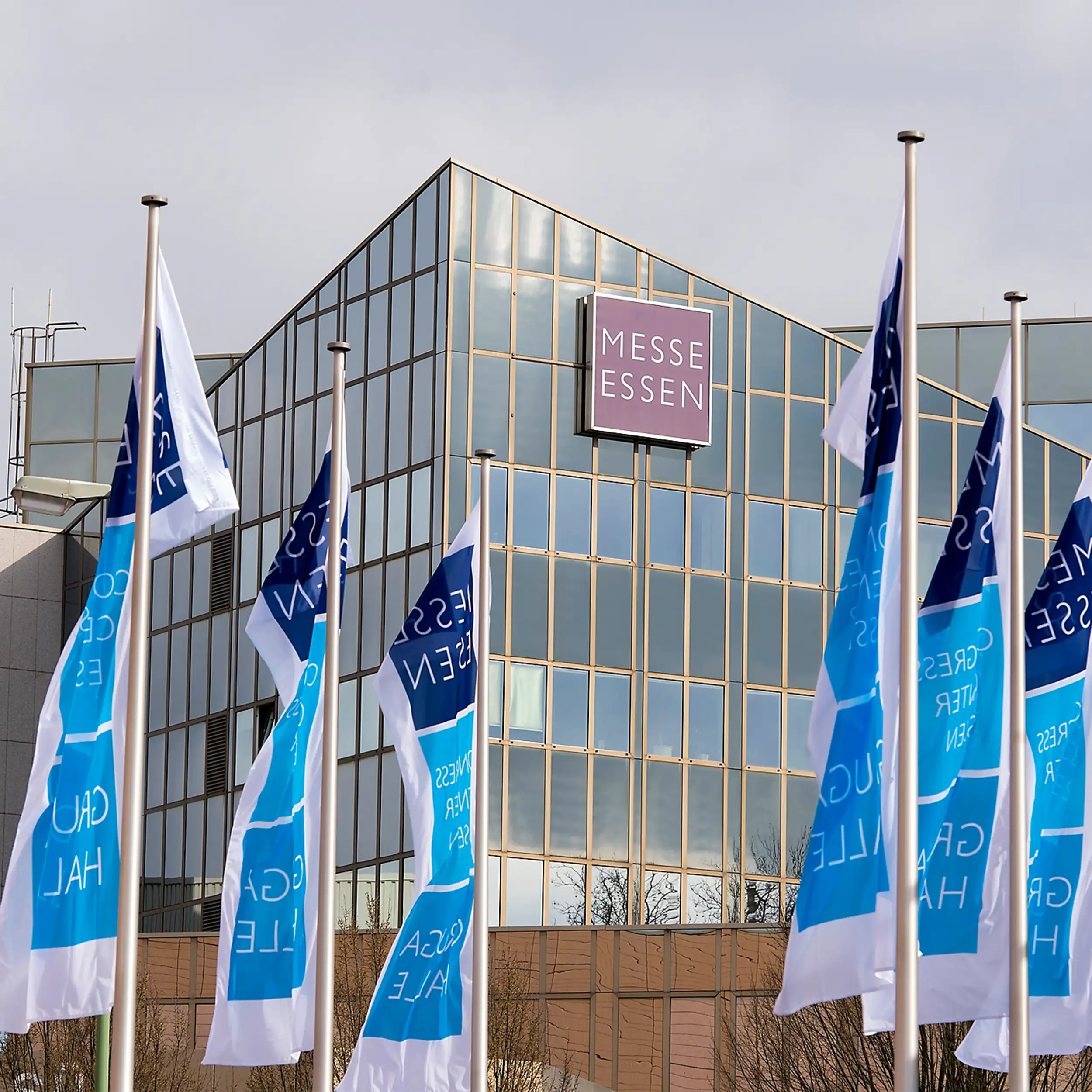 Außenansicht der Messe Essen mit Glasfassade, Messe-Essen-Schild und mehreren Fahnen im Vordergrund.