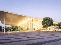 Beleuchteter Eingangsbereich der Messe Essen mit moderner Glasfassade, großem Vordach und Bäumen im Vordergrund.