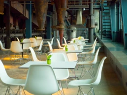 Innenbereich des Café Kohlenwäsche mit weißen Stühlen und Tischen vor historischer Industriekulisse.