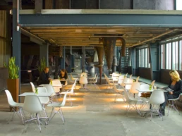 Café Kohlenwäsche mit weißen Tischen und Stühlen, industrieller Architektur und mehreren Gästen im Innenbereich.